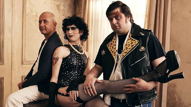 A bald man in a black blazer sits next to a person dressed as Dr. Frank-N-Furter from "Rocky Horror Picture Show" and another cosplayer.