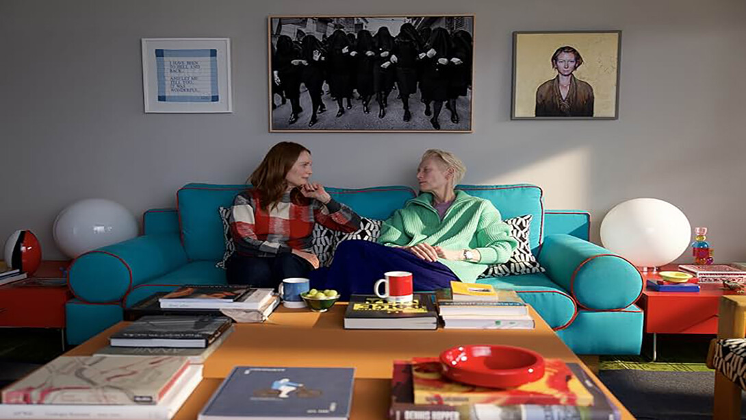 A redheaded woman in a red and white flannel shirt sits to the left of a blonde woman in a green robe on a turquoise couch.