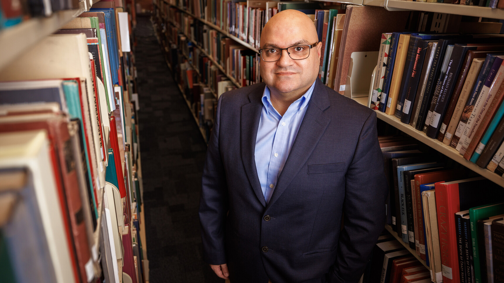 Bedross Der Matossian, Hymen Rosen Professor in Judaic Studies at the University of Nebraska–Lincoln, stands between rows of bookshelves in Love Library. He is wearing a dark blue sport coat over a light blue dress shirt, and glasses.